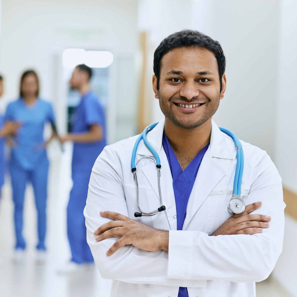 Portrait of experienced doctor from India with crossed arms and stethoscope standing in hallway of modern hospital. Medical profession